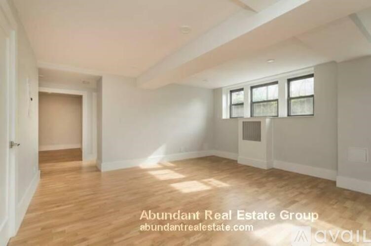 A spacious room with wooden flooring and white walls, with a door on the left and a window with blinds on the right.