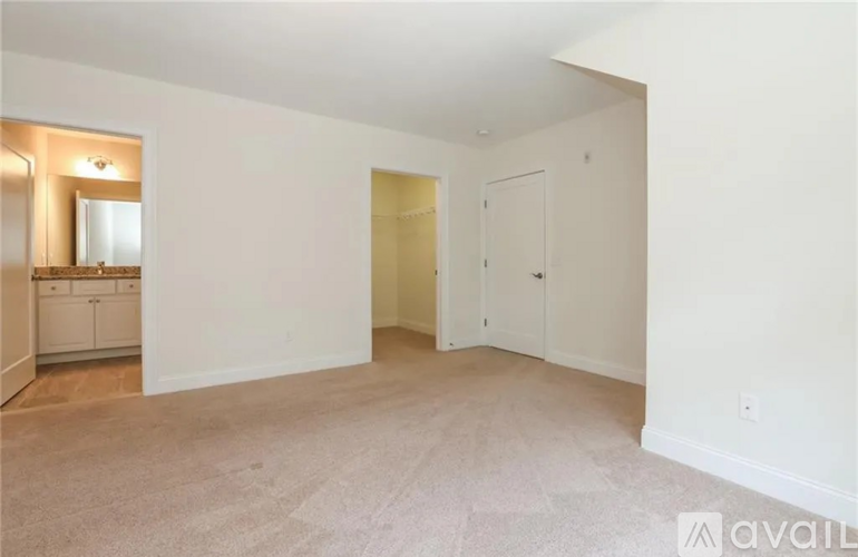 A spacious room with beige carpeting and white walls, featuring a bathroom with a vanity and mirror, a door, and a window.