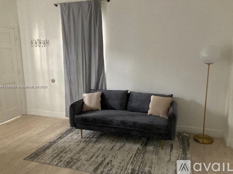 A black sofa with two pillows sits in a room with a grey curtain and a wooden floor.