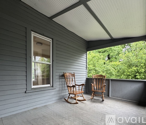 Two rocking chairs on a porch with a window and trees in the background.