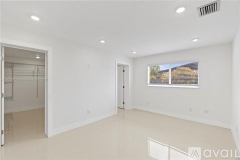 A spacious, empty room with white walls and flooring, a window with blinds, and a door leading to another room.