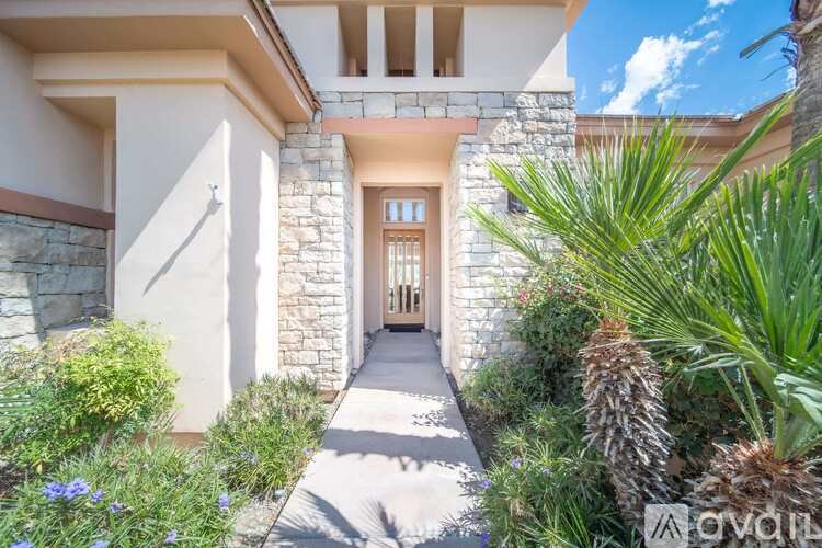 A house with a stone wall and a wooden door is surrounded by green plants.