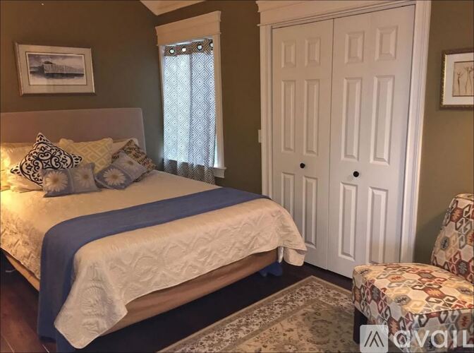 A bedroom with a bed, a window with curtains, a white door, and a patterned ottoman.