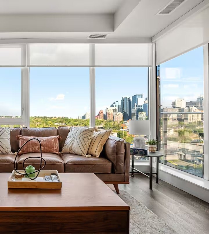 A brown leather couch with a wooden tray on top of it in front of a window with a city view.