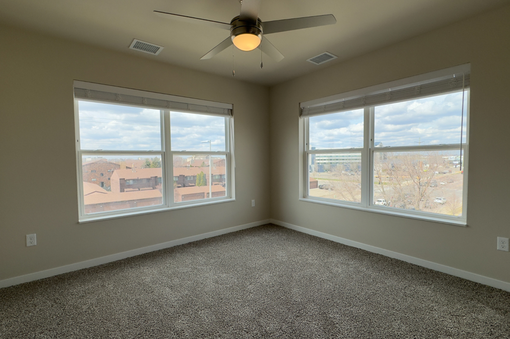 A room with a ceiling fan and two windows overlooking a residential area.