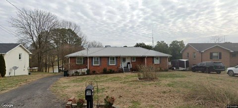 A house with a red brick exterior and a grey roof is surrounded by a grassy yard and a dirt path leading to a mailbox.