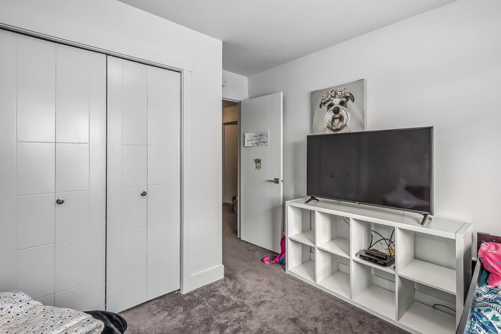 A room with a television on a stand and a bed with a white comforter.