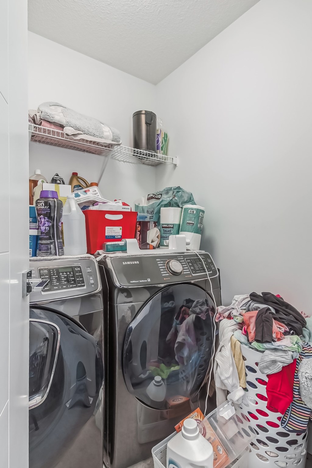 A laundry room with a washer and dryer, a basket of clothes, and a shelf with various items.