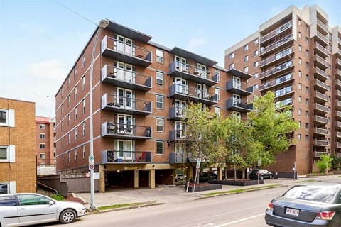 A street view of a residential area with cars parked on the side of the road and apartment buildings in the background.