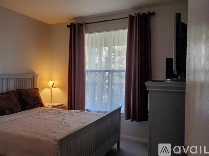 A bedroom with a bed, a nightstand with a lamp, a window with curtains, and a television.