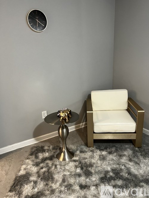 A chair with a gold base and a white cushion sits in front of a grey wall with a clock on it.