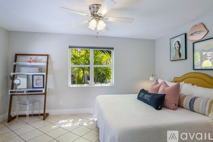 A bedroom with a bed, a window with a view of greenery, a ceiling fan, and a picture frame on a stand.