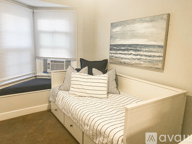 A bedroom with a bed, a window with blinds, and a painting of a beach scene on the wall.