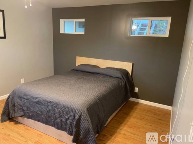 A bed with a grey comforter is in a room with wooden floors and grey walls.