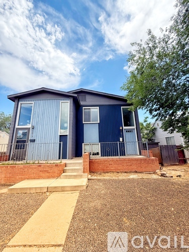 A blue house with a white door and windows is surrounded by a gravel driveway and a brick wall.