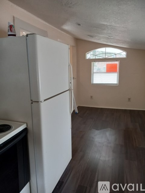 A white refrigerator stands in a room with wood flooring and a window with blinds.