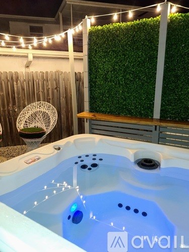 A hot tub with lights around it is in the foreground with a green hedge and wooden fence in the background.
