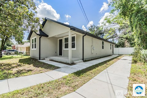 A small house with a front porch is surrounded by greenery and a sidewalk.