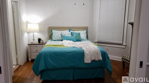 A bed with a teal comforter and a white blanket is in the foreground of a room with a lamp and a nightstand.