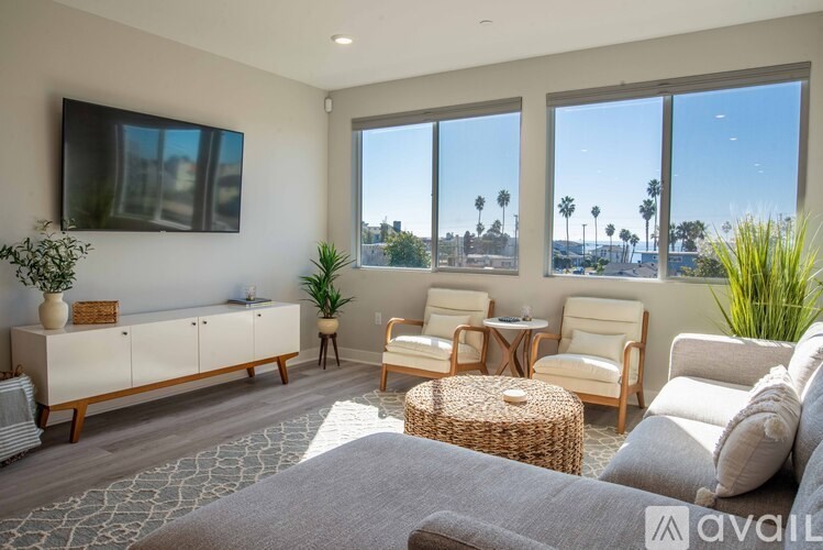 A living room with a grey rug, a grey sofa, a wooden cabinet, a TV, a table with chairs, and a window with a view of palm trees.