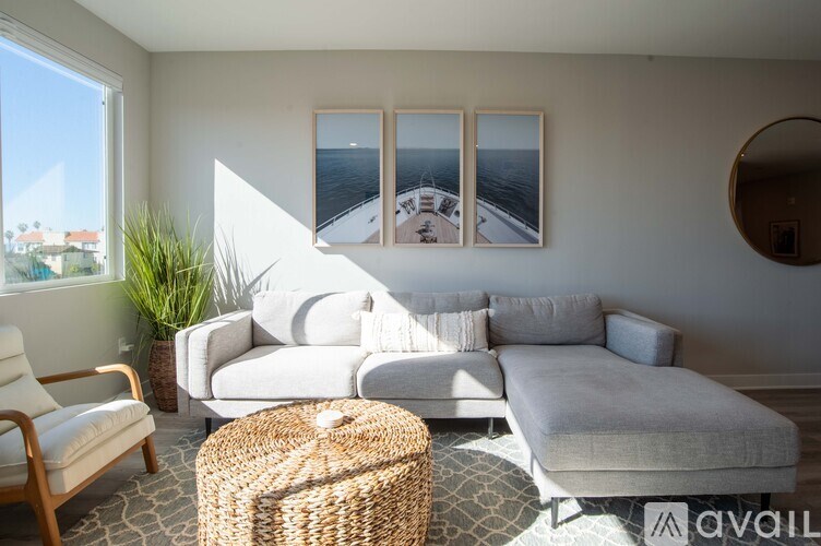 A living room with a white couch, a wooden chair, a wicker ottoman, and a large window with a view of the city.