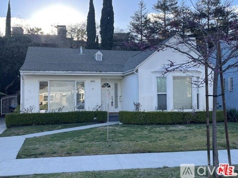 A white house with a grey roof and a chimney is surrounded by greenery.
