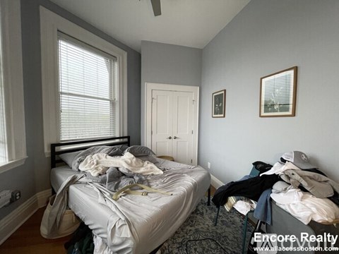 A bedroom with a bed in the center of the room with a grey bedspread and a chair with clothes on it.