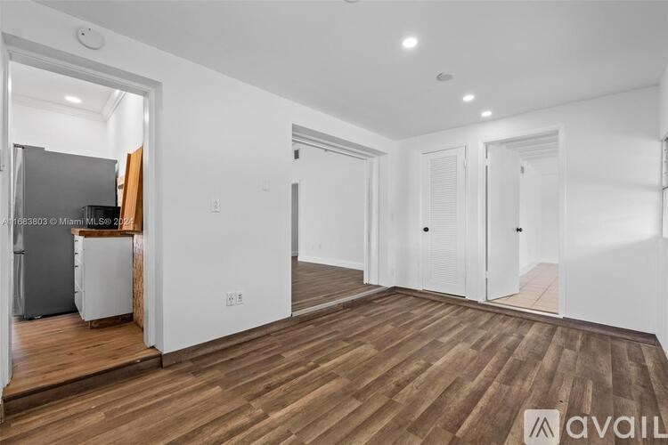 A room with wooden flooring and white walls, with a kitchen visible through the open door.