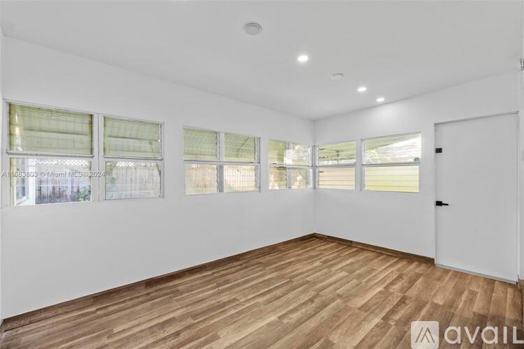 A room with wooden flooring and white walls, with a door on the right and windows on the left.