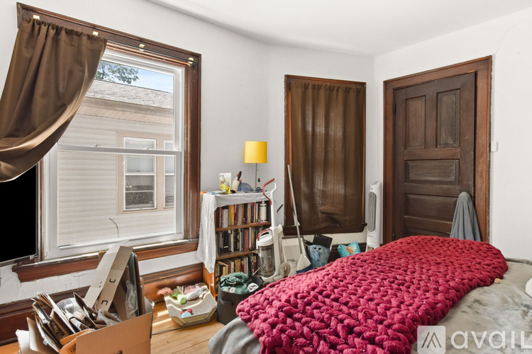 A bedroom with a bed covered in a red blanket, a window with brown curtains, and a wooden door.