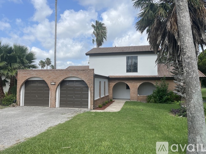 A house with a white exterior and a grey roof is surrounded by greenery and palm trees.