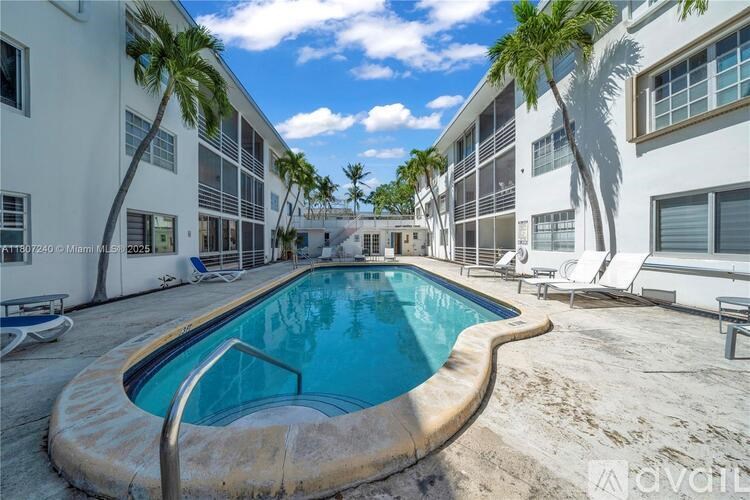 A swimming pool in the middle of a sandy area with palm trees and white buildings in the background.