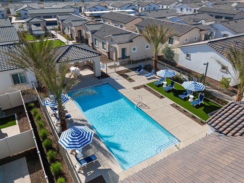 A swimming pool surrounded by white lounge chairs with blue cushions and blue and white striped umbrellas with a green area at Allero 59  Apartments, Phoenix, Arizona