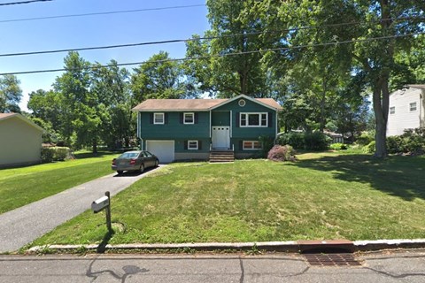 A green house with a white door and windows is surrounded by trees and a car is parked in the driveway.