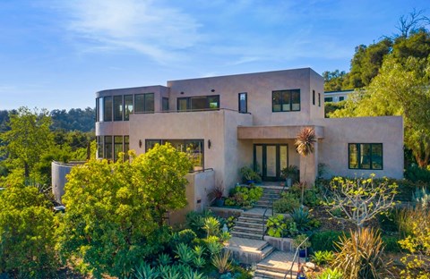 A modern house with a flat roof and large windows is surrounded by greenery.
