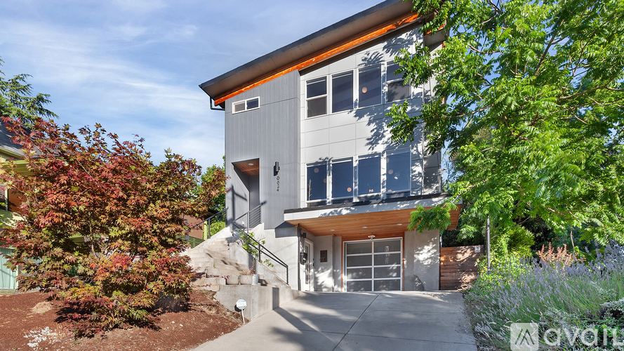 A modern house with a grey exterior and a brown roof is surrounded by greenery and has a sign that says "available".