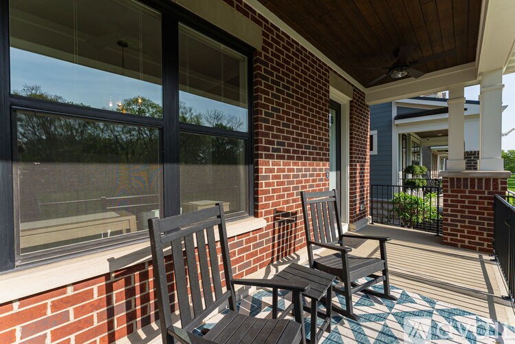 A wooden chair sits on a porch with a brick wall and a window.