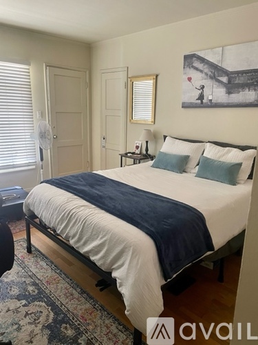 A bedroom with a bed, a fan, a rug, a painting, and a small table.