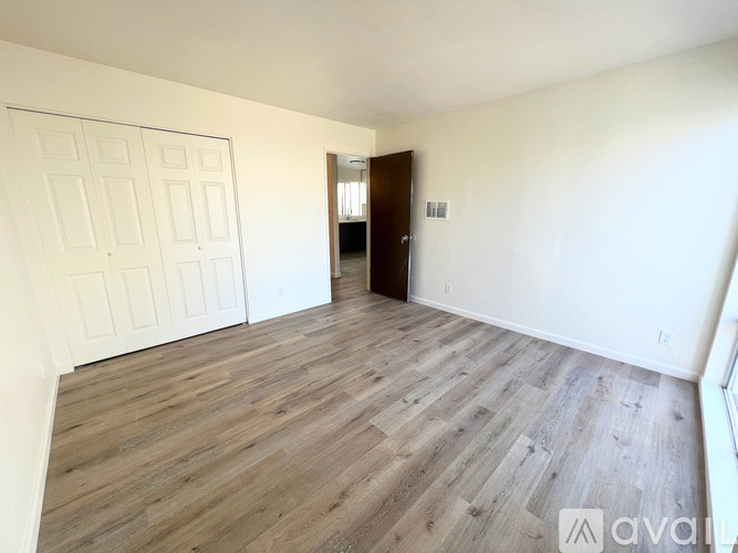 A room with wooden flooring and white walls, with a door on the left and a window on the right.