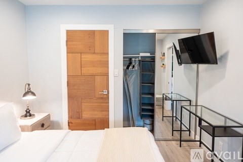 A bedroom with a bed, a nightstand with a lamp, a wooden door, a staircase, a glass railing, and a flat-screen TV mounted on the wall.