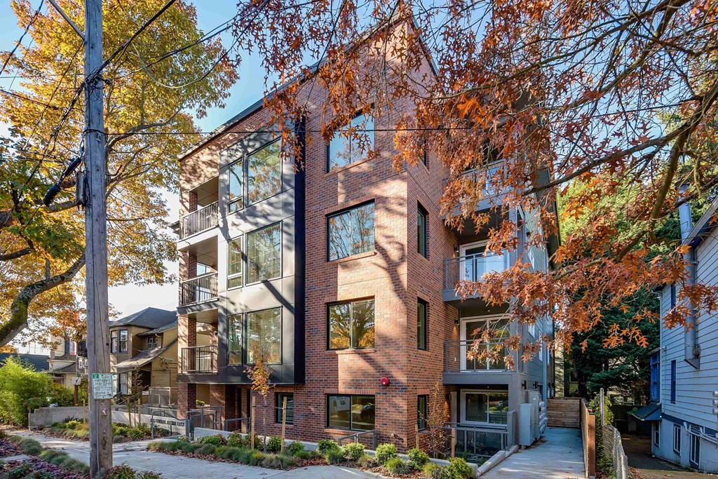 A modern brick building with glass windows and balconies is surrounded by trees with autumn leaves.