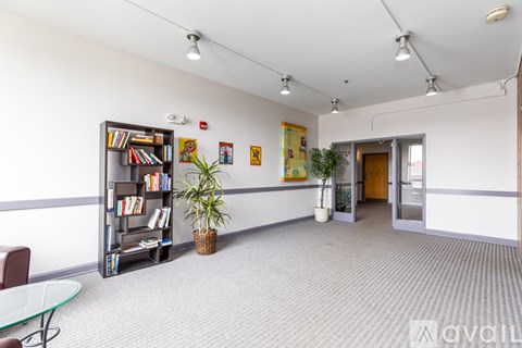 A room with a carpeted floor, a glass table, a bookshelf, and a few potted plants.
