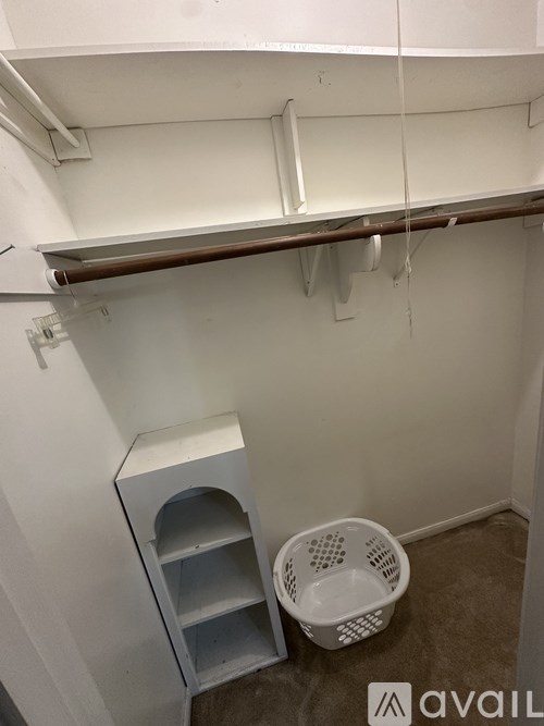 A white laundry basket sits on a carpeted floor next to a white shelf unit.