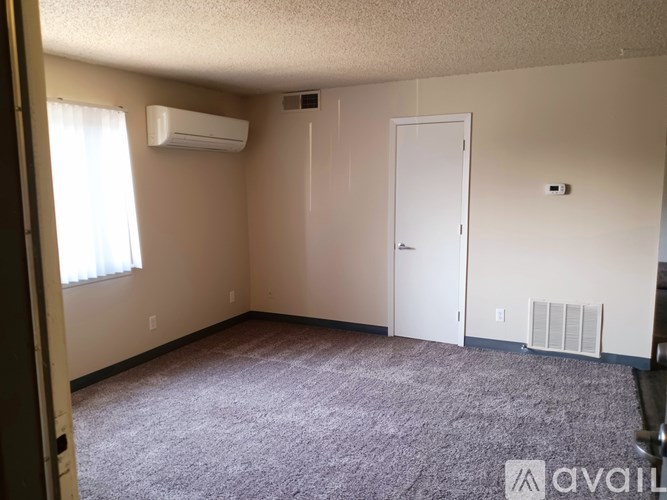 A room with a carpeted floor, a window with white curtains, a white door, and two air conditioning units on the wall.