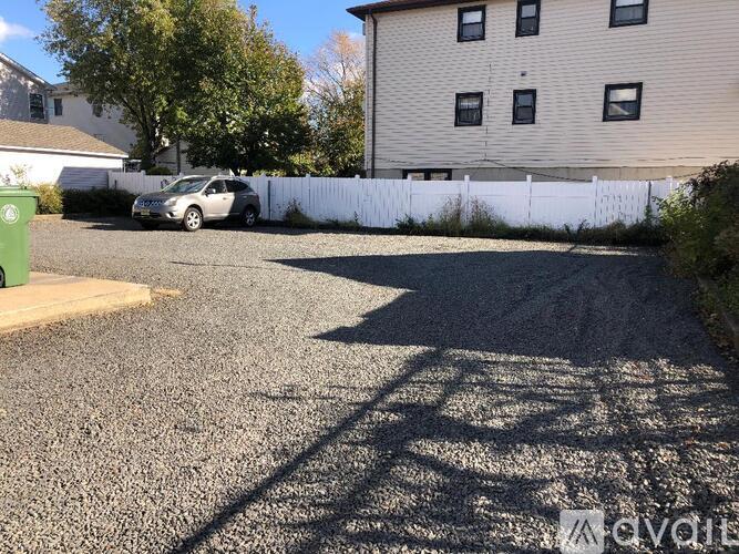 A car is parked in a gravel driveway in front of a white fence and apartment building.