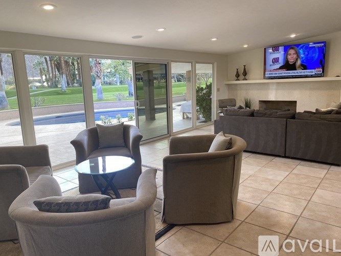 A living room with a television on the wall and a view of a pool and trees outside the window.