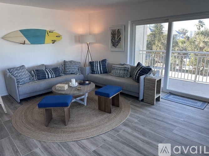 A living room with a grey couch, a blue ottoman, a coffee table, and a surfboard on the wall.