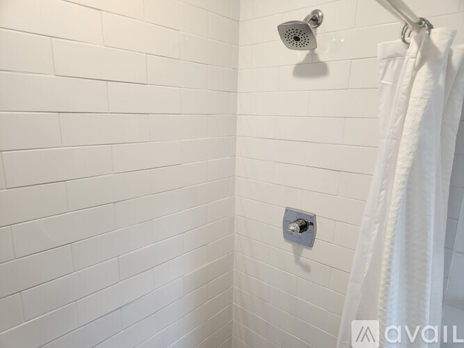 A white tiled bathroom wall with a shower head and a white towel hanging on a towel rack.