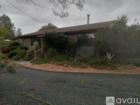 A house with a brown roof and a chimney is surrounded by greenery and a gravel driveway.