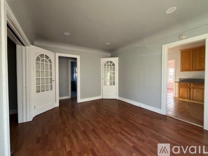A room with wooden floors and white walls, with a kitchen visible in the background.
